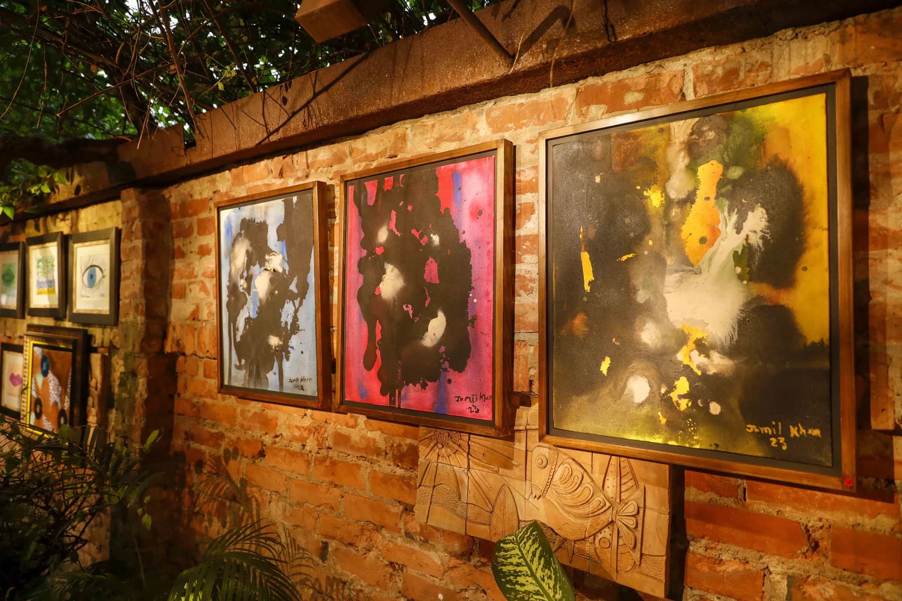 Framed paintings by Jamil Khan hung along a lit brick garden wall